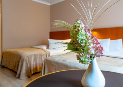 Nahaufnahme eines Nachttischs mit Vase, frischen Blumen, im Hintergrund zwei Betten.