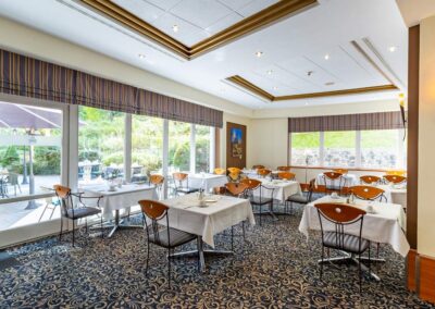 Restaurantbereich mit gedeckten Tischen, großen Fenstern und blauem Teppichboden mit Rankenmuster.