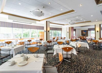 Restaurantbereich mit gedeckten Tischen, großen Fenstern und blauem Teppichboden mit Rankenmuster.